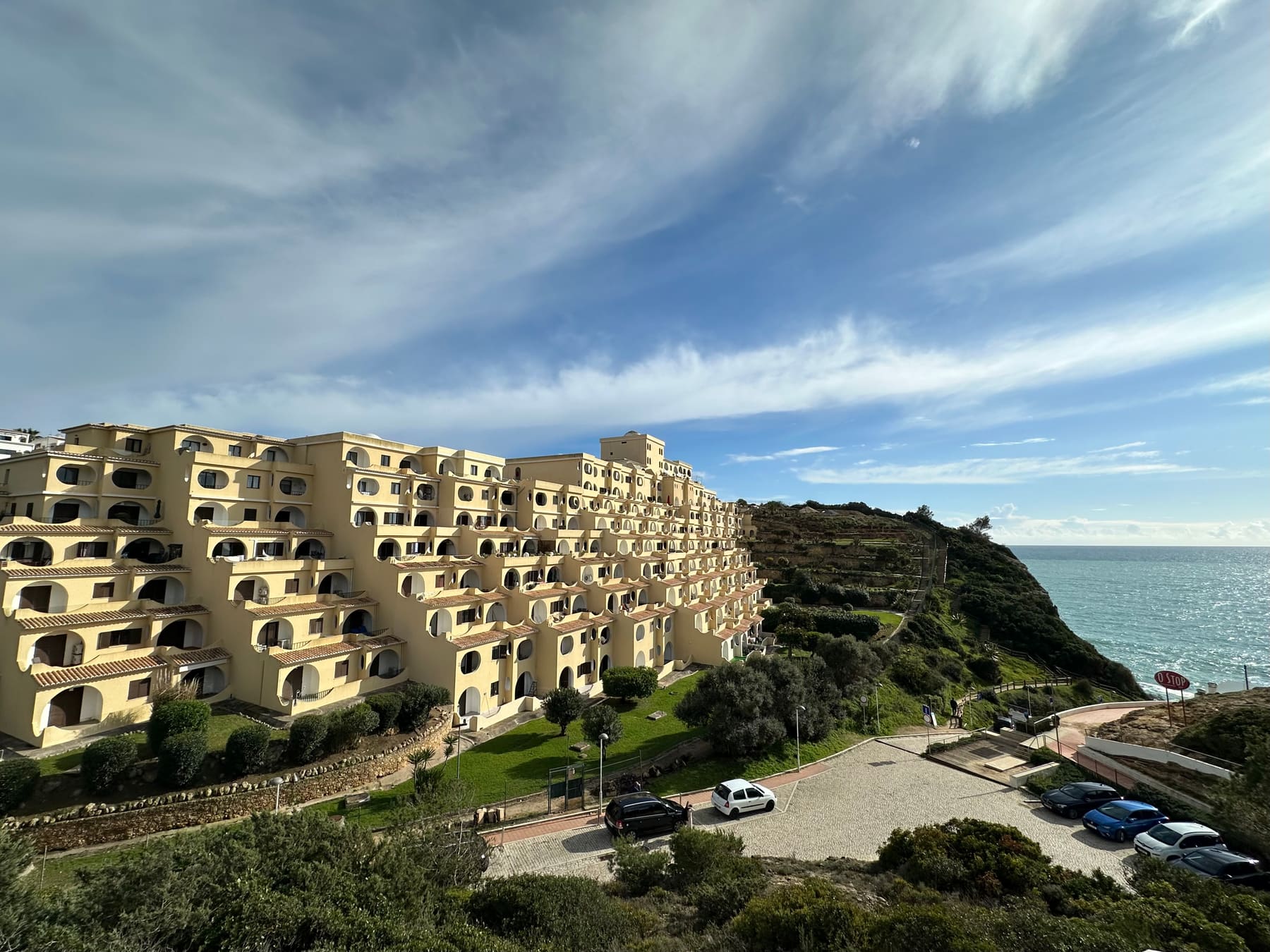 Terraced apartment and hotel buildings on the clifftop above the sea at Vale Centeanes, with a road and parked cars below.